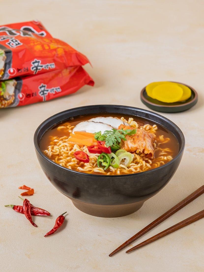A bowl of Shin Ramyun instant noodles topped with mushrooms, carrots, kimchi, green onion and red chili, with two Shin Ramyun packages behind it