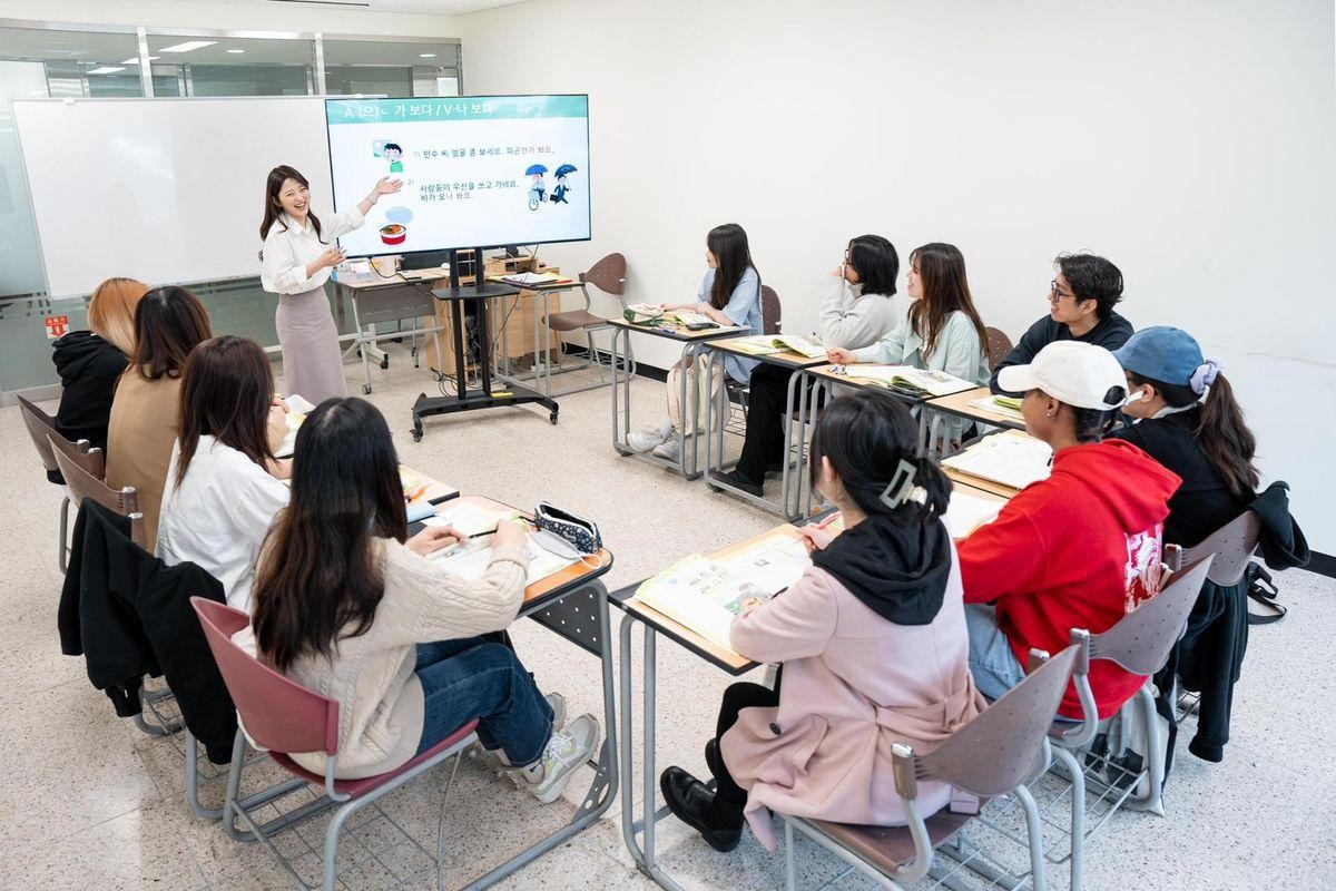 A classroom scene with a teacher standing by a screen and students sitting at desks.