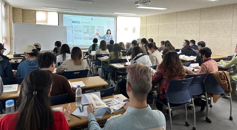 A woman stands at the front of a larger classroom, presenting to a group of students seated at desks.