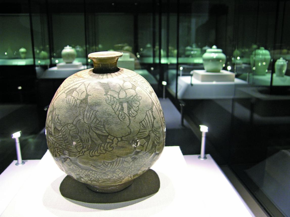 A celadon-glazed vessel with inlaid floral and fish motifs displayed in a museum gallery