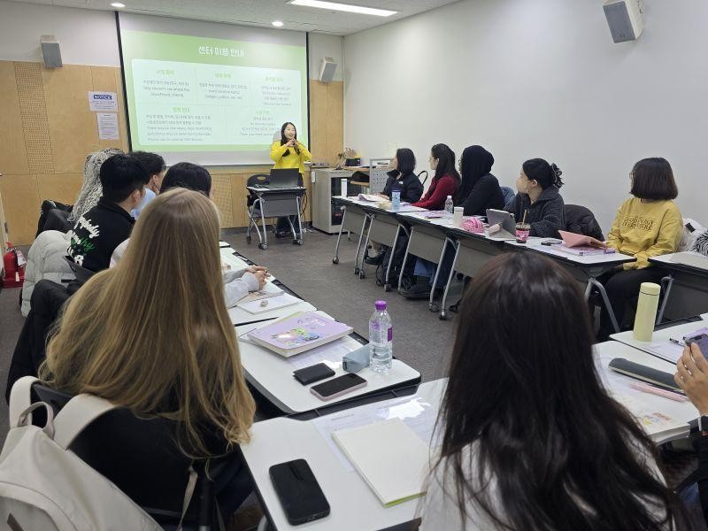 A woman in a yellow shirt stands at the front of a classroom, presenting to a group of students seated at desks.