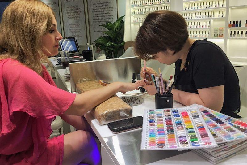 International visitor having her nails done by a Korean nail artist beside a large gel colour chart in a Seoul salon