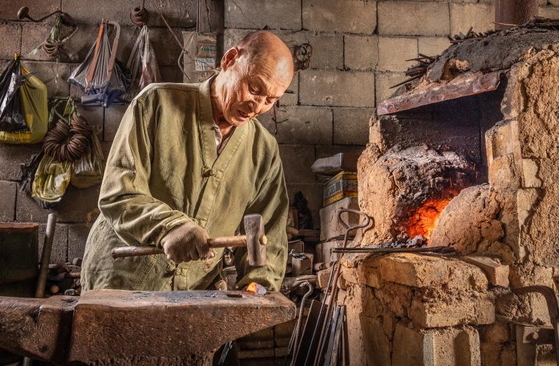 A Korean blacksmith hammering a piece of glowing iron on an anvil beside the forge